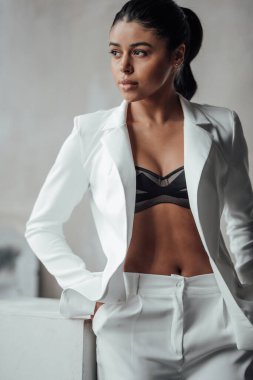 Genç, yetişkin, esmer bir kadın resmi bir gece elbisesi ve düşünceli bir sütyenle. Şık, kıvırcık saçlı, şehvetli, Afro-Amerikan manken modayla stüdyoda moda kıyafeti içinde poz veriyor.