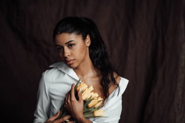 Beyaz gömlekli, esmer, genç ve güzel bir kadın çıplak omuzlarıyla sarı lale buketini kucaklıyor. Siyah kıvırcık saçlı, şehvetli Afro-Amerikan model moda uzmanı bahar tatilinde stüdyoda poz veriyor.