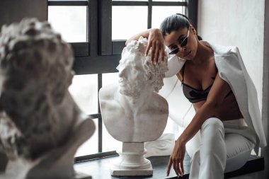 Beyaz takım elbiseli, esmer, genç bir kadın. Tişörtsüz, Yunan erkeği büstü heykeli. Şık, kıvırcık saçlı, şehvetli, Afro-Amerikan manken modayla stüdyoda moda kıyafeti içinde poz veriyor.