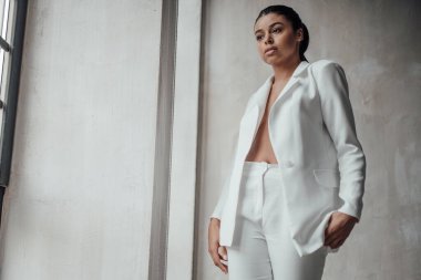 Genç, yetişkin, esmer bir kadın resmi bir gece elbisesi ve düşünceli bir sütyenle. Şık, kıvırcık saçlı, şehvetli, Afro-Amerikan manken modayla stüdyoda moda kıyafeti içinde poz veriyor.