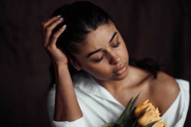 Beyaz gömlekli, esmer, genç ve güzel bir kadın çıplak omuzlarıyla sarı lale buketini kucaklıyor. Siyah kıvırcık saçlı, şehvetli Afro-Amerikan model moda uzmanı bahar tatilinde stüdyoda poz veriyor.
