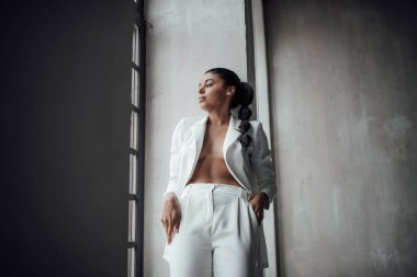 Genç, yetişkin, esmer bir kadın resmi bir gece elbisesi ve düşünceli bir sütyenle. Şık, kıvırcık saçlı, şehvetli, Afro-Amerikan manken modayla stüdyoda moda kıyafeti içinde poz veriyor.