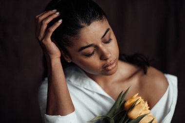 Beyaz gömlekli, esmer, genç ve güzel bir kadın çıplak omuzlarıyla sarı lale buketini kucaklıyor. Siyah kıvırcık saçlı, şehvetli Afro-Amerikan model moda uzmanı bahar tatilinde stüdyoda poz veriyor.