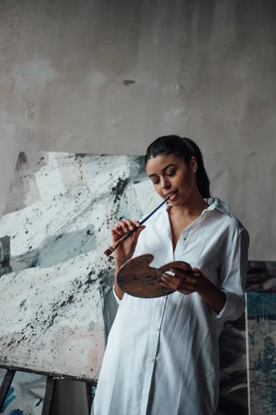 年轻的艺术家 身穿白色长衫的黑衣女子 光着腿站在她的绘画上 画室里有时尚的黑色卷发性感的非洲美式模特 配上画板和画笔 — 图库照片