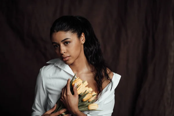 Beyaz gömlekli, esmer, genç ve güzel bir kadın çıplak omuzlarıyla sarı lale buketini kucaklıyor. Siyah kıvırcık saçlı, şehvetli Afro-Amerikan model moda uzmanı bahar tatilinde stüdyoda poz veriyor.