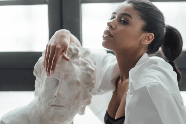 stock image Young beauty swarthy woman in formal evening white suit no shirt hugs greek man bust statue. Stylish black curly hair sensual african american model fashionista posing at studio in fashion pantsuit