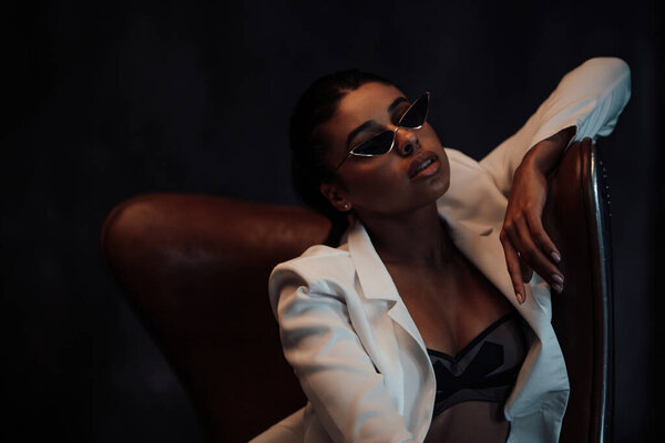 Young adult beauty swarthy woman in formal evening white suit with bra at cat eye sunglasses. Stylish black curly hair sensual african american model fashionista posing at studio in fashion pantsuit