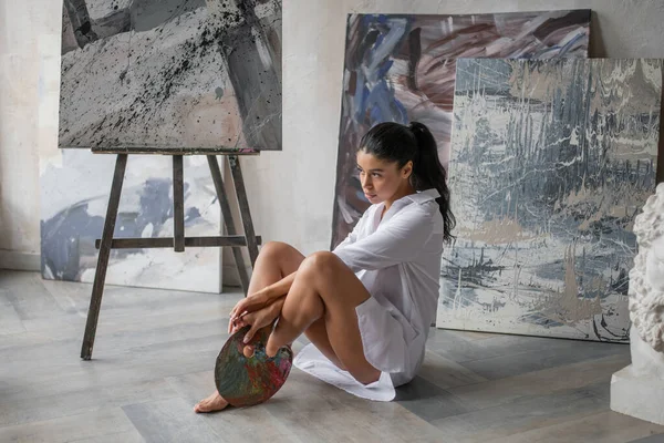 stock image Young artist beauty swarthy woman in white long shirt bare legs posing against her paintings. Stylish black curly hair sensual african american model with paint palette and brushes at painting studio