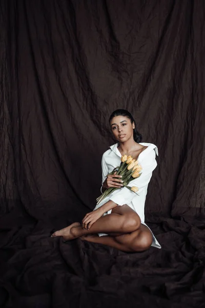 stock image Young adult beauty swarthy woman in white shirt bare shoulder hugs bouquet of yellow tulips. Stylish black curly hair sensual african american model fashionista posing in studio at spring holidays