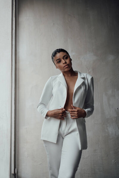 Young adult beauty swarthy woman in formal evening white suit with bra at thoughtful. Stylish black curly hair sensual african american model fashionista posing at studio in fashion pantsuit