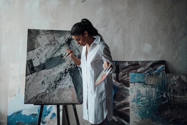 年轻的艺术家 身穿白色长衫的黑衣女子 光着腿站在她的绘画上 画室里有时尚的黑色卷发性感的非洲美式模特 配上画板和画笔 — 图库照片
