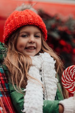 Noel 'i ve yeni yılı kutlayan bayan çocuk kış tatilini dışarıda kutluyor. Kırmızı örgü şapkalı aktif küçük kız elinde Noel şekeri bastonuyla açık havada vakit geçiriyor.