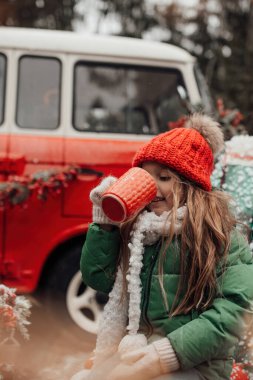 Noel 'i ve yeni yılı kutlayan bayan çocuk kış tatilini dışarıda kutluyor. Kırmızı örülmüş şapkalı küçük bir kız açık havada vakit geçirip Xmas otobüsünün yanındaki büyük bardaktan sıcak çikolata içiyor.