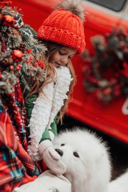 Noel 'i ve yeni yılı kutlayan bayan çocuk kış tatilini dışarıda kutluyor. Küçük kız açık havada beyaz Samoylu köpekle Noel otobüsünün yanında eğleniyor. Çocukluğun tadını çıkarıyor.