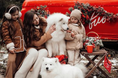 Mutlu aile Noel 'i ve yeni yılı dışarıda kutluyor. Anneleri ve beyaz Samoyed köpekleri olan çocuklar birlikte vakit geçirip eski Xmas otobüsünün yanında sarılıyorlar.