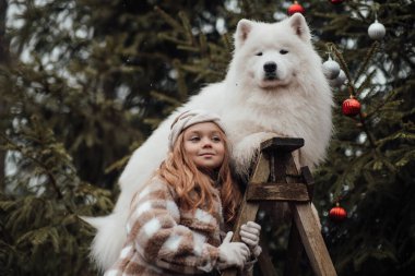 Noel 'i ve yeni yılı kutlayan bayan çocuk kış tatilini dışarıda kutluyor. Küçük kız açık havada beyaz Samoylu köpekle Noel otobüsünün yanında eğleniyor. Çocukluğun tadını çıkarıyor.