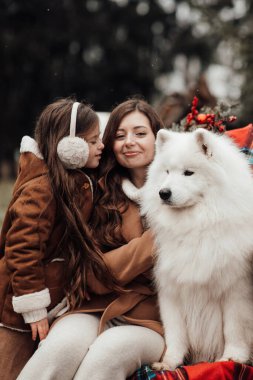 Mutlu aile Noel 'i ve yeni yılı dışarıda kutluyor. Annesi ve beyaz Samoyed köpeği olan kız birlikte vakit geçirip eski Xmas otobüsünün yanında sarılıyor.