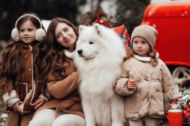 Mutlu aile Noel 'i ve yeni yılı dışarıda kutluyor. Anneleri ve beyaz Samoyed köpekleri olan çocuklar birlikte vakit geçirip eski Xmas otobüsünün yanında sarılıyorlar.