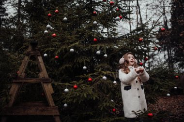 Noel 'i ve yeni yılı kutlayan bayan çocuk kış tatilini dışarıda kutluyor. Aktif küçük kız kozalaklı ormanda çocukluğunun tadını çıkarıyor.