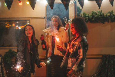 Mutlu kızlar Noel 'i ve yeni yıl tatilini dışarıda kutluyor. Aktif, genç, çeşitli kadınlar neşeli bayan arkadaşlar ve Bengal ışıkları maytapları eski Xmas karavanının yanında eğleniyorlar.