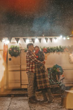 Noel 'i ve yeni yılı kutlayan mutlu çift, kış tatili sezonunu Camper Park' ta kutluyor. Genç ve neşeli çift birlikte vakit geçiriyorlar. Kucaklaşıp öpüşüyorlar. Yılbaşı karavanı yakınlarında ilk kar yağışına seviniyorlar.