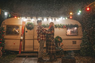 Noel 'i ve yeni yılı kutlayan mutlu çift, kış tatili sezonunu Camper Park' ta kutluyor. Genç ve neşeli çift birlikte vakit geçiriyorlar. Kucaklaşıp öpüşüyorlar. Yılbaşı karavanı yakınlarında ilk kar yağışına seviniyorlar.