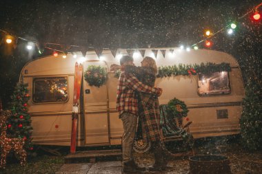 Noel 'i ve yeni yılı kutlayan mutlu çift, kış tatili sezonunu Camper Park' ta kutluyor. Genç ve neşeli çift birlikte vakit geçiriyorlar. Kucaklaşıp öpüşüyorlar. Yılbaşı karavanı yakınlarında ilk kar yağışına seviniyorlar.
