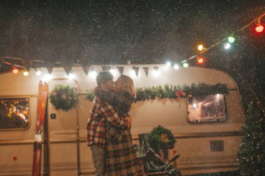 Noel 'i ve yeni yılı kutlayan mutlu çift, kış tatili sezonunu Camper Park' ta kutluyor. Genç ve neşeli çift birlikte vakit geçiriyorlar. Kucaklaşıp öpüşüyorlar. Yılbaşı karavanı yakınlarında ilk kar yağışına seviniyorlar.