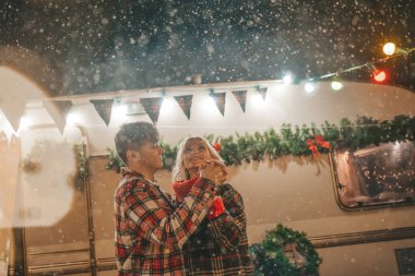 Noel 'i ve yeni yılı kutlayan mutlu çift, kış tatili sezonunu Camper Park' ta kutluyor. Genç ve neşeli çift birlikte vakit geçiriyorlar. Kucaklaşıp öpüşüyorlar. Yılbaşı karavanı yakınlarında ilk kar yağışına seviniyorlar.