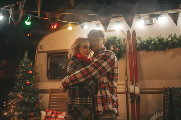 Happy couple celebrating Christmas and New Year winter holidays season in Camper Park. Young couple rest and relaxation spending time together hugs and kisses near Xmas camper trailer