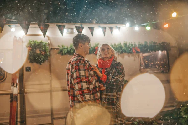 Happy couple celebrating Christmas and New Year winter holidays season in Camper Park. Young joyful couple spending time together hugs and kisses rejoices at first snow near Xmas camper trailer