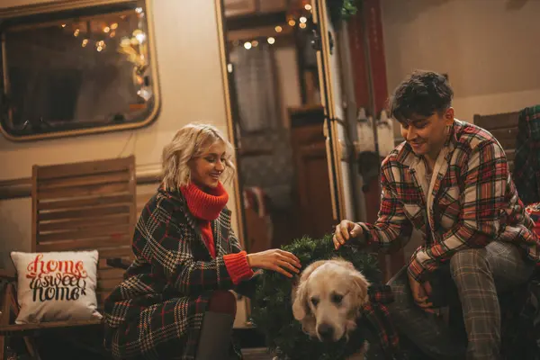 Happy couple celebrating Christmas and New Year winter holidays season in Camper Park. Young joyful couple hugging and spending time together with golden retriever dog near Xmas camper trailer