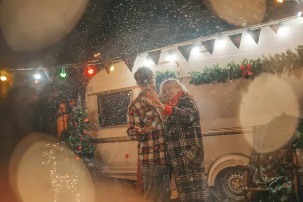 Happy couple celebrating Christmas and New Year winter holidays season in Camper Park. Young joyful couple spending time together hugs and kisses rejoices at first snow near Xmas camper trailer
