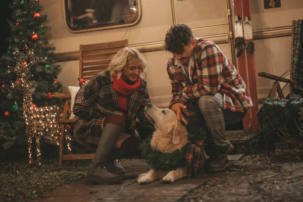 Happy couple celebrating Christmas and New Year winter holidays season in Camper Park. Young joyful couple hugging and spending time together with golden retriever dog near Xmas camper trailer