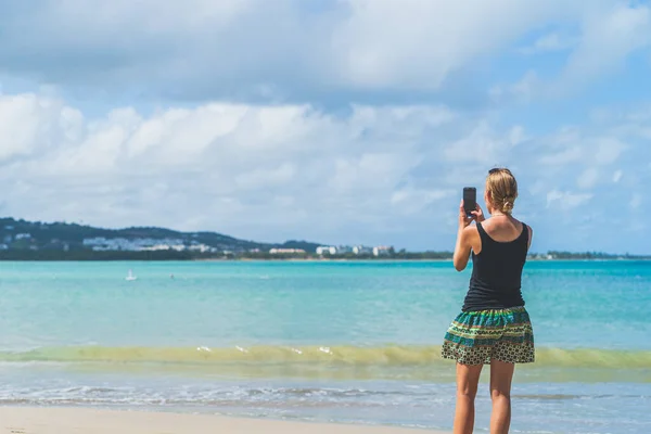 stock image Woman using phone camera on beach with smartphone mobile phone against teal waters and clouds.