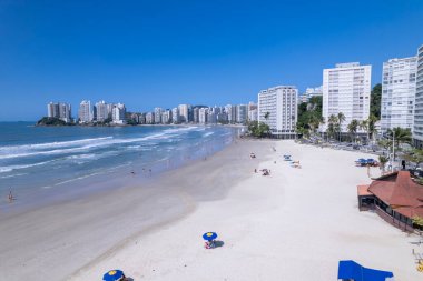 Bu kıyı manzarasında Guaruja, Sao Paulo 'daki Praia da Pitangueiras, açık mavi suların altın kumlarla buluştuğu ve kıyı boyunca uzanan yüksek binalar ziyaretçileri plajın tadını çıkarmaya davet ediyor..