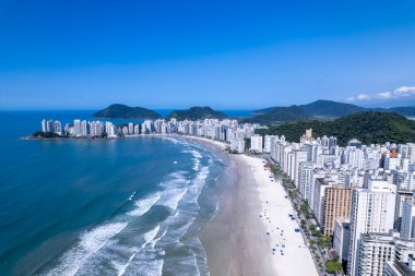 Bu kıyı manzarasında Guaruja, Sao Paulo 'daki Praia da Pitangueiras, açık mavi suların altın kumlarla buluştuğu ve kıyı boyunca uzanan yüksek binalar ziyaretçileri plajın tadını çıkarmaya davet ediyor..