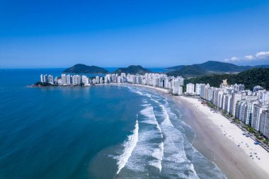 Bu kıyı manzarasında Guaruja, Sao Paulo 'daki Praia da Pitangueiras, açık mavi suların altın kumlarla buluştuğu ve kıyı boyunca uzanan yüksek binalar ziyaretçileri plajın tadını çıkarmaya davet ediyor..