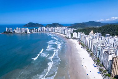 Bu kıyı manzarasında Guaruja, Sao Paulo 'daki Praia da Pitangueiras, açık mavi suların altın kumlarla buluştuğu ve kıyı boyunca uzanan yüksek binalar ziyaretçileri plajın tadını çıkarmaya davet ediyor..
