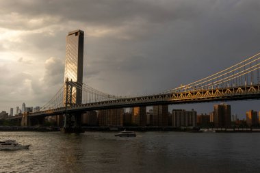 Alacakaranlık New York 'a inerken Williamsburg Köprüsü Doğu Nehri boyunca uzanır ve altında gemiler seyreder. Ufuk çizgisi sıcak yansımalarla parlıyor..