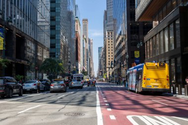 New York 'ta hareketli bir şehir caddesi, parlak mavi gökyüzünün altında yükselen gökdelenlerle çevrili araçlarla dolu. İnsanlar kaldırımlarda gezinir, canlı atmosferin tadını çıkarırlar..
