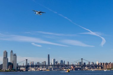 Bir hidrojen uçağı New York siluetinin üzerinde geziniyor ve şehrin kendine özgü mimarisini açık mavi gökyüzü zeminine karşı sergiliyor..