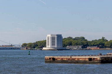New York 'un su kıyısında, ağaçlarla çevrili tarihi bir yapı ve mavi bir gökyüzü altında sakin bir liman, kentsel huzuru ön plana çıkarıyor..