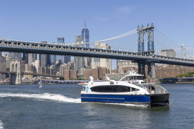 Bir feribot, New York 'un ufuk çizgisi görünen Manhattan Köprüsü' nün altından geçerek Doğu Nehri 'ni geçer. Parlak mavi gökyüzü sahneyi büyütüyor..