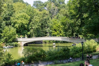 Aydınlık bir günde, insanlar teknelerde dinleniyor ve Central Park 'taki büyüleyici bir taş köprüde yürüyor, ağaçlarla çevrili ve sakin bir gölet, doğanın şehirdeki güzelliğini vurguluyor..