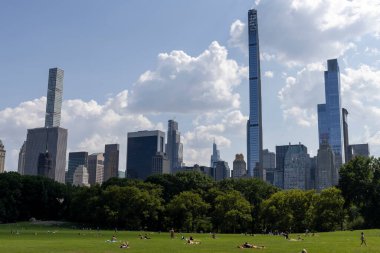 Parlak bir gökyüzü altında ziyaretçiler New York City parkının yemyeşil çimlerinde dinlenirken modern gökdelenler arka planda görkemli bir şekilde yükseliyor ve kentsel yaşamı gözler önüne seriyorlar..