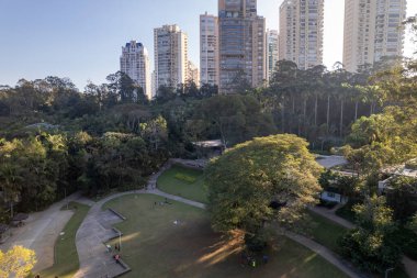 Sao Paulo 'daki yüksek gökdelenlerin arasına kurulmuş geniş bir şehir parkı, güneşli bir öğleden sonra açık havada eğlenen insanlarla dolu yemyeşil ve patikalarla dolu..