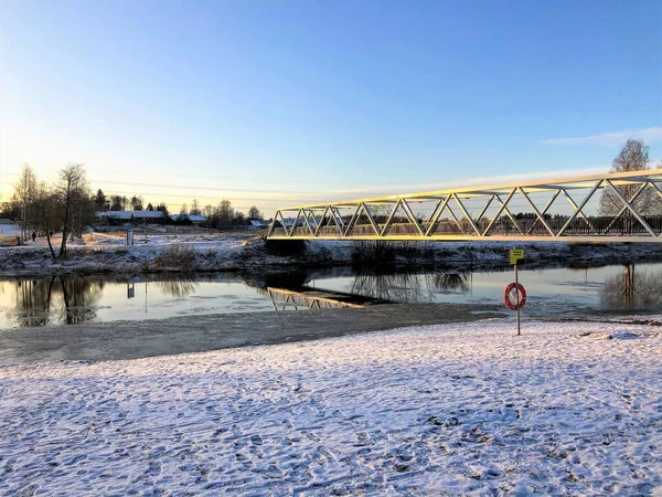 Iron geometrical bridge over the partly frozen river with life buoy ans snow surface with many foot prints and trees and buildings on the background lighted by low winter sun