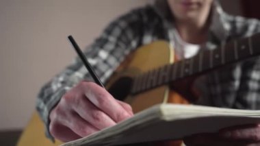 Genç adam rahat bir odada akustik gitarla şarkı sözleri yazıyor. yakın plan
