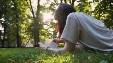 Genç ve çekici bir kız yeşil çimlerde uzanıyor ve kitap okuyor..
